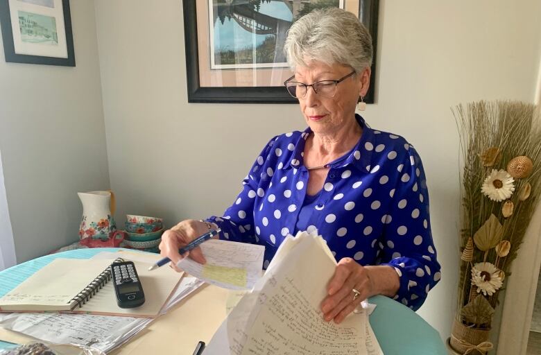 An elderly woman wearing a blue blouse sorts through hand-written notes strewn across her kitchen table.