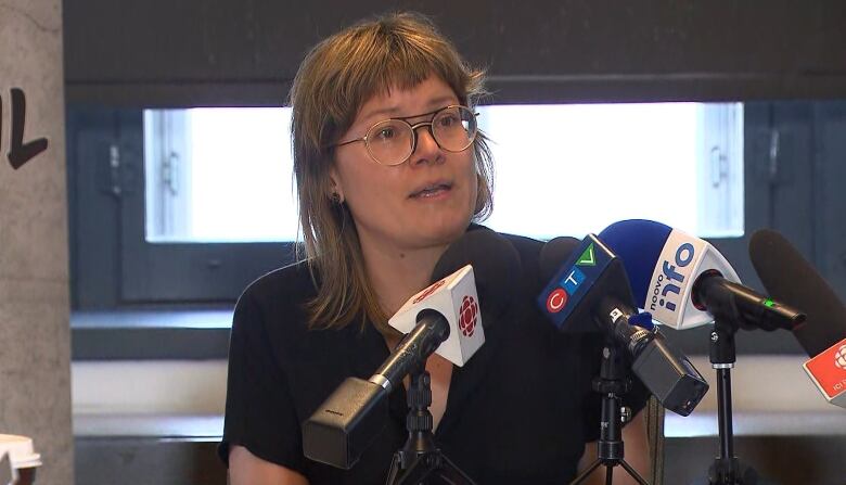 A white woman with short bangs and glasses speaks into a set of media microphones in front of  window.