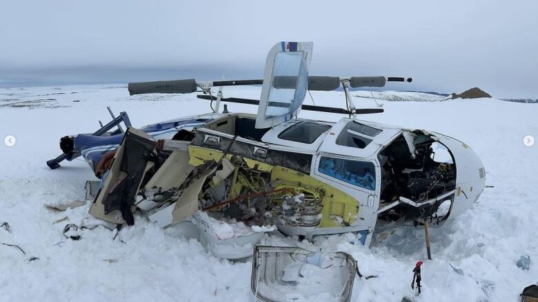 The wreckage of a crashed helicopter lies on its side on an icy, snowy expanse.