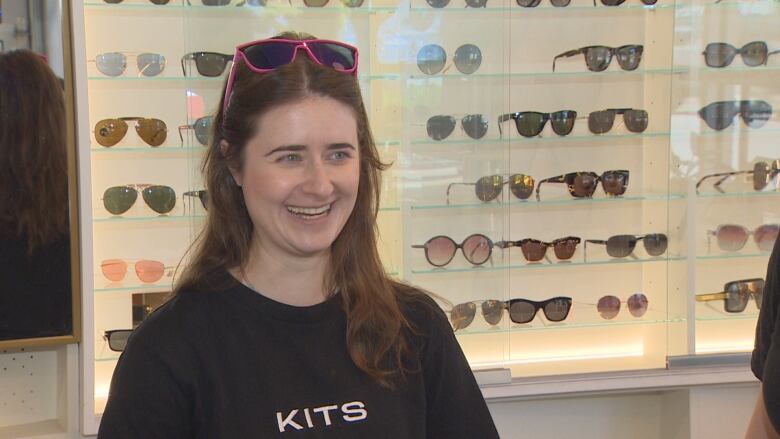 A woman with long brown hair and blue eyes stands behind a shelf of sunglasses.