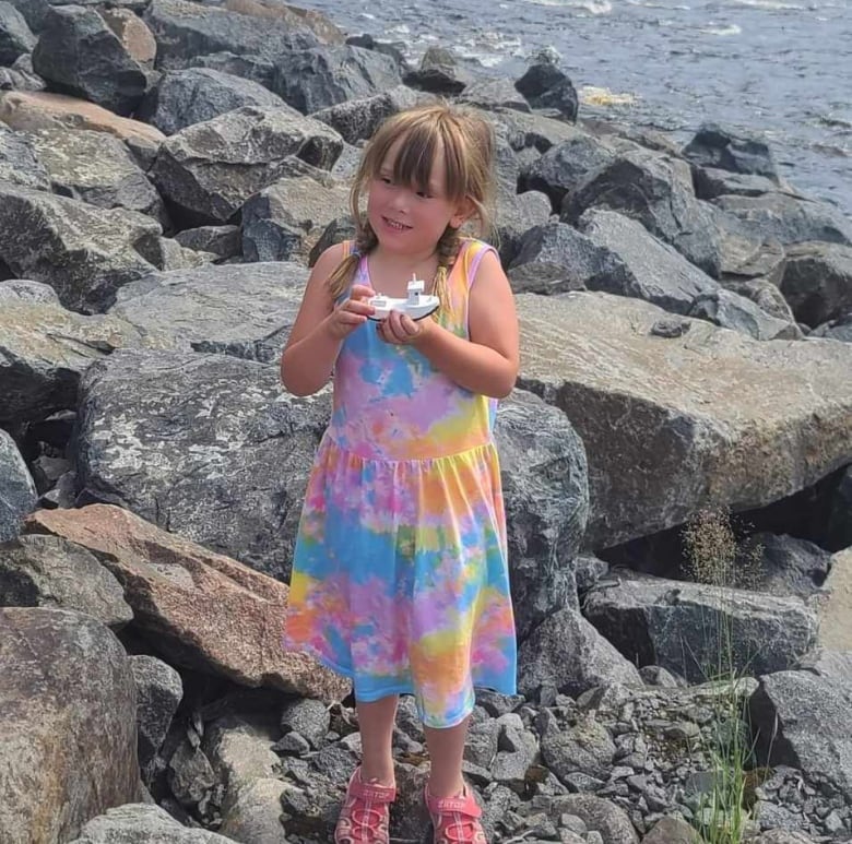 A girl in a rainbow dress holds a boat while standing on rocks. 