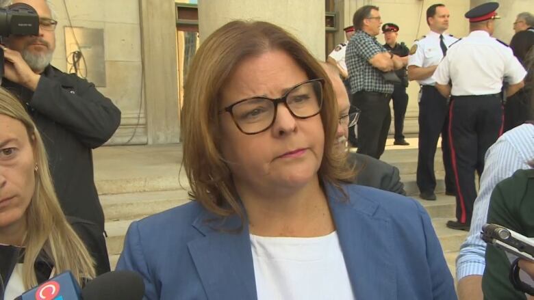 A woman wearing glasses and a blue blazer speaks to reporters holding microphones.