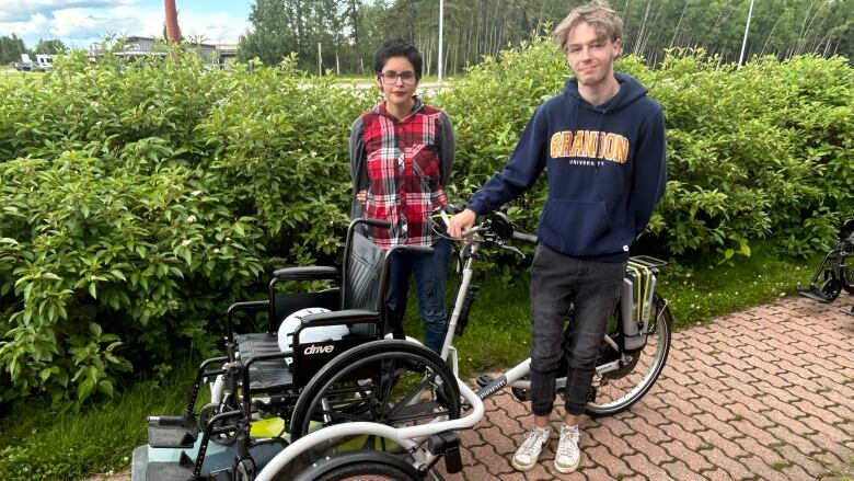 A wheelchair and two teens standing behind it. 