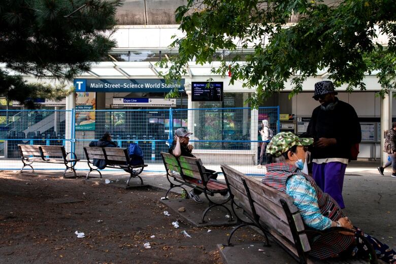 Pedestrians and passersby are pictured seated on benches, walking in front of the Main Street-Science World SkyTrain Station, whose entrance is bordered with a blue fence.