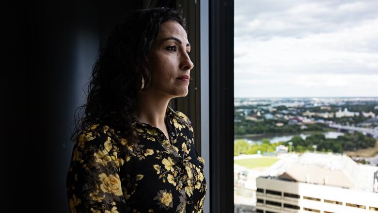 A woman stands beside a window and looks out at a view of downtown Winnipeg.