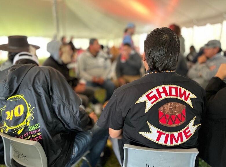 Two men in a crowded tent sit with their backs to the camera.