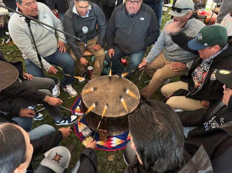 A circle of men sit banging on a drum.