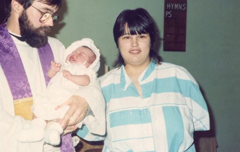 An old photograph shows a priest holding a baby with a woman smiling beside them. 