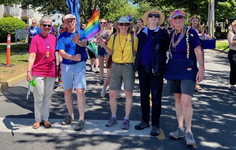 Five people in colourful clothing stand together, with a person behind raising a rainbow flag.