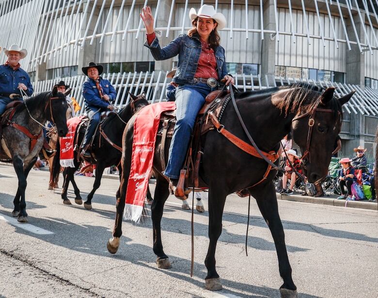 a woman in a blue jean jacket and red blouse rides a horse on a city street and waves