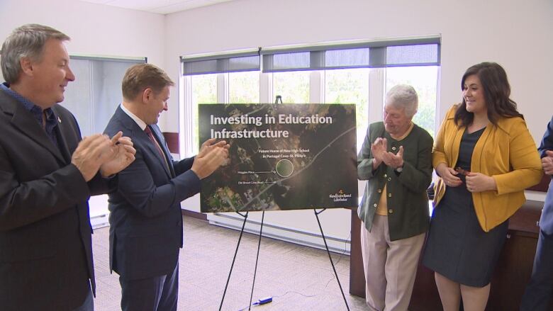 Four clapping people stand next to a poster board. The board reads 'Investing in Education Infrastructure' and shows the location where a new high school will be built.