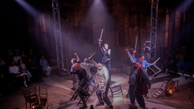 A group of actors, shouting, brandish chairs and wooden swords off stage on a round circle as audience members look on.