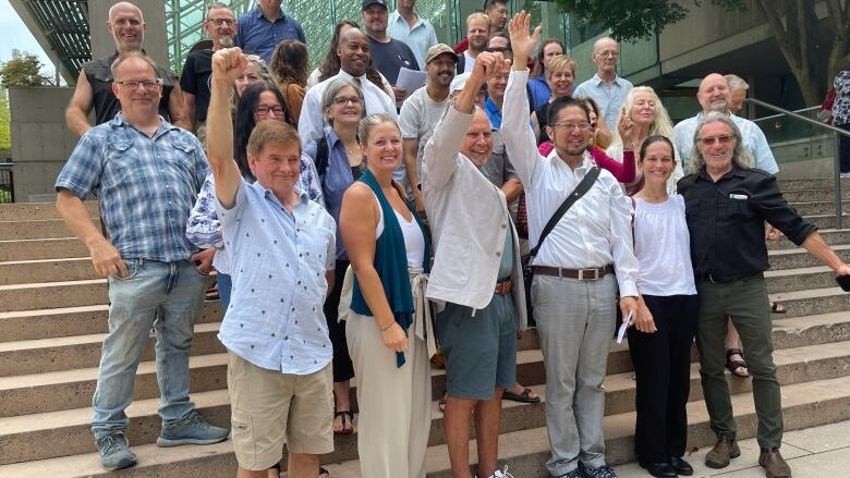 A group of about two dozen people stand in five rows on concrete steps outside a court building. They are smiling and raising their fists in the air.