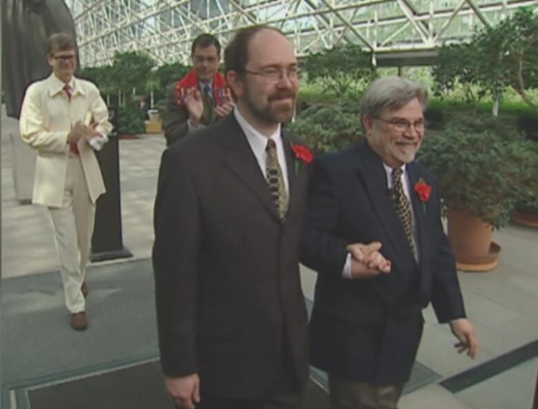 Two men are pictured in suits, holding hands, with red rose corsage. They are smiling, walking with a minister behind them. 