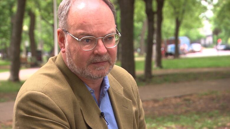 A white man sitting at a table outside wearing a brown blazer and glasses with grey hair.