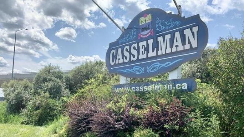 A town sign on a summer day, surrounded by greenery.