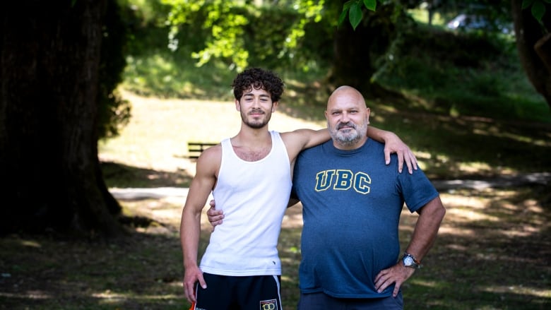 A young man in his late teens and a man in his fifties stand arm-in-arm in a wooded park