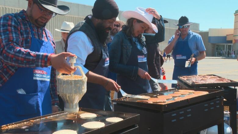 A crowd of people stand around a griddle with pancakes in a parking lot. One of them is a woman in a cowboy hat. 