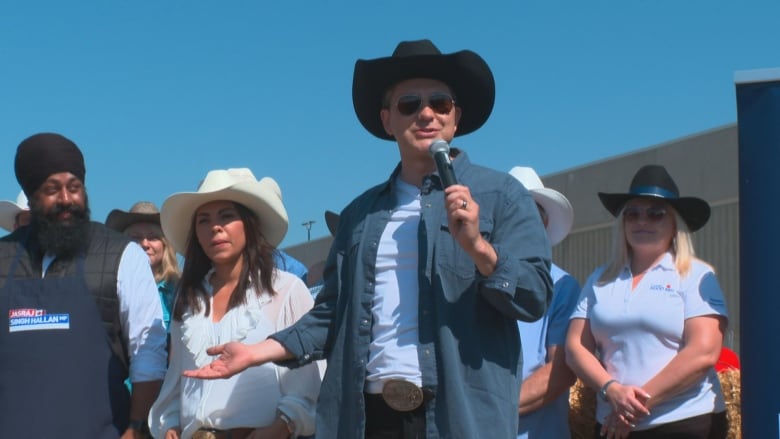 A man in a cowboy hat speaks into a microphone. There are people standing around him. 