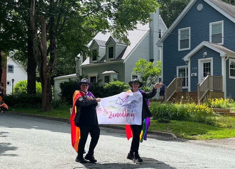 Two people draped in pride flags hold a banner saying Lunenburg.