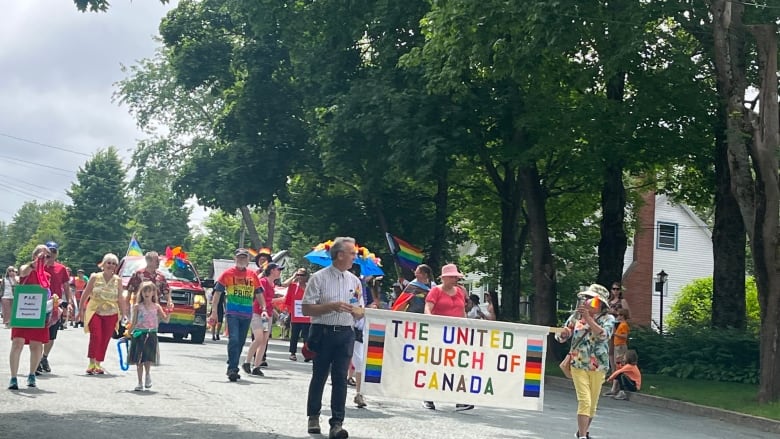 People with pride flags and colours walk on a street led by two people holding a banner saying United Church of Canada.