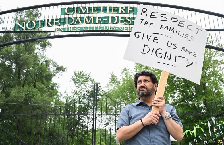 Jimmy Koliakoudakis holds up a sign during a protest outside Notre-Dame-des-Neiges Cemetery in Montreal, Sunday, July 9, 2023. The sign reads: Respect the families, give us some dignity.