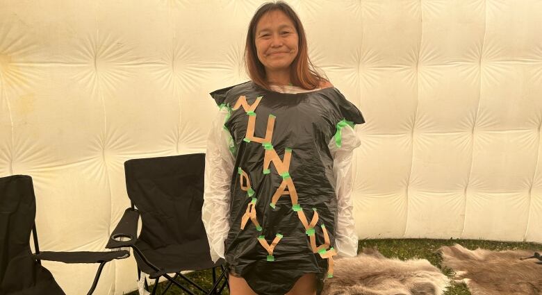 A woman smiles to the camera. She's wearing a makeshift dress from a black garbage bag, which has cardboard letters on them that say 