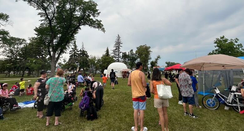 A group of people are gathered on a field. Some are sitting, and some stand. An inflatable igloo is seen in the distance.