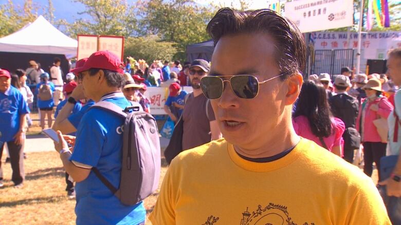 An East Asian man talks to a camera while wearing a yellow shirt at an outdoor event.