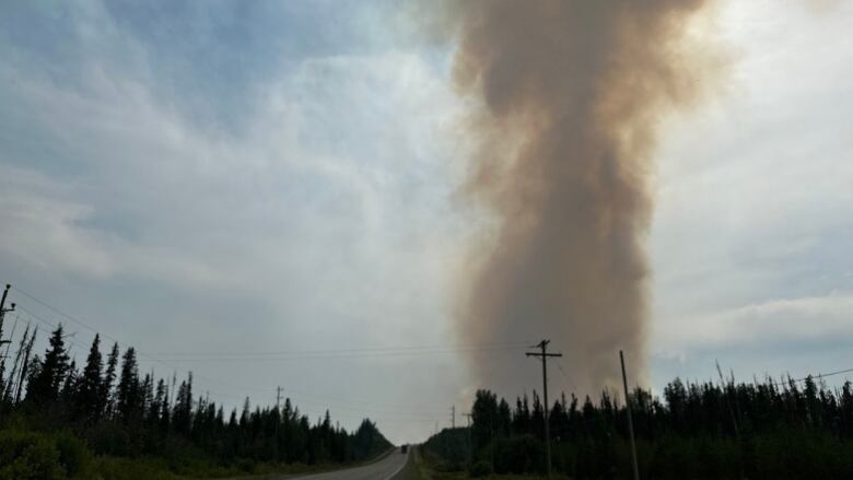 Smoke rises turning the sun red in the distance, with a highway and forest in the foreground