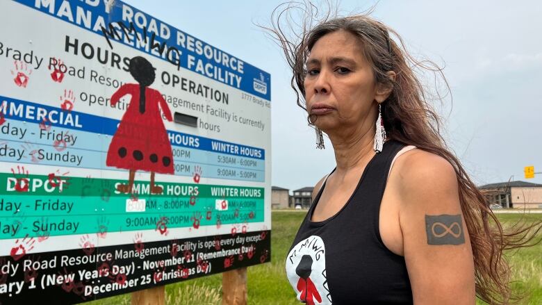 A woman stands next to a landfill hours of operation sign. Red handprints and a woman in a red dress are painted on top.