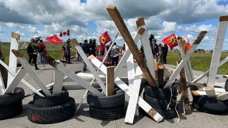 Planks of wood standing upright inside of car tires line a road creating a blockade. People are on the other side of the blockade with red and yellow flags.