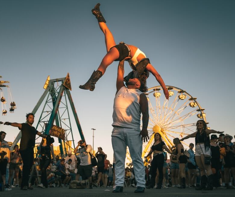 A man stands on the Stampede Grounds with one arm raise as his dance partner is balanced on his hand about him in an arch with one leg kicking up in the air.