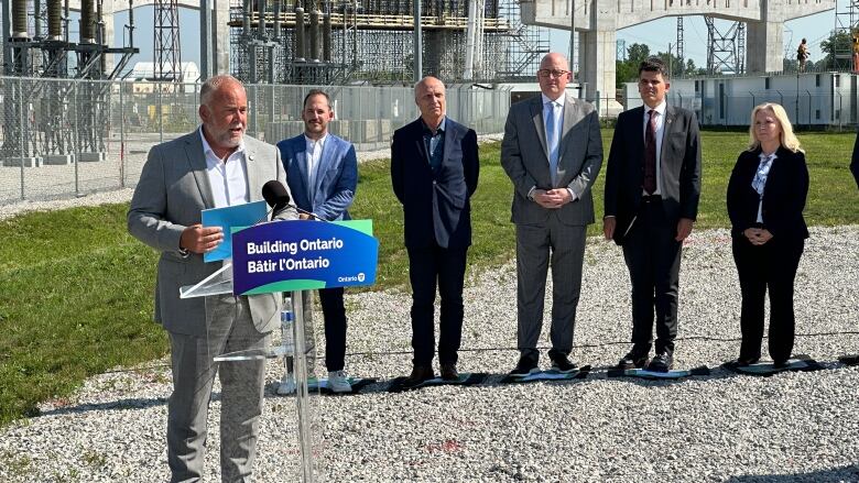 A man speaks at a podium in front of a power plant, four other men and one woman stand behind him .