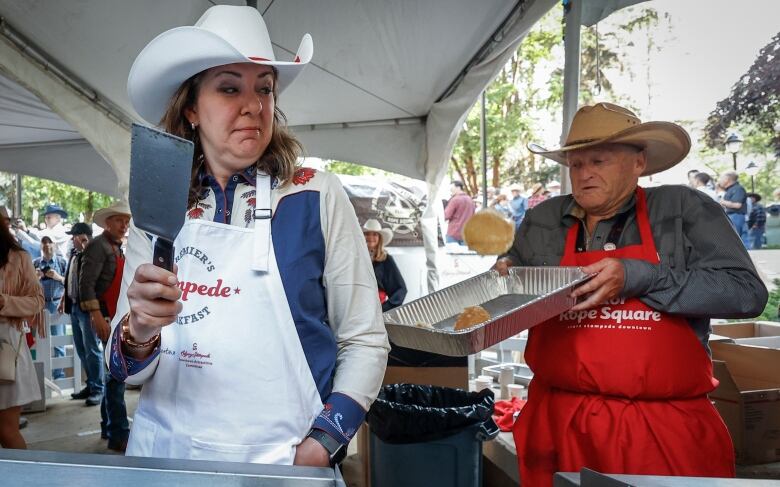 A woman wearing a cowboy hat holds a spatula as she flips a pancake over her shoulder. A man holding tray moves to catch a pancake in the pan.