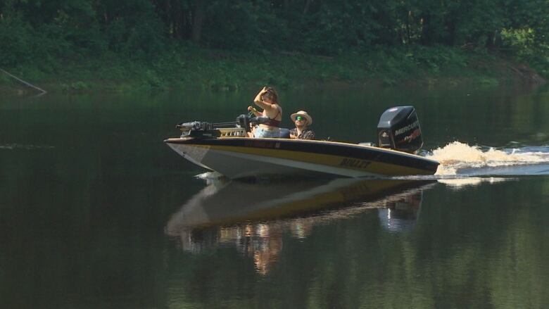 A yellow boat drives up the Nashwaak river. Two people sit inside.