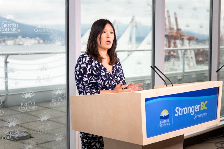 An East Asian woman speaks at a podium while a port and white sails are seen behind her.