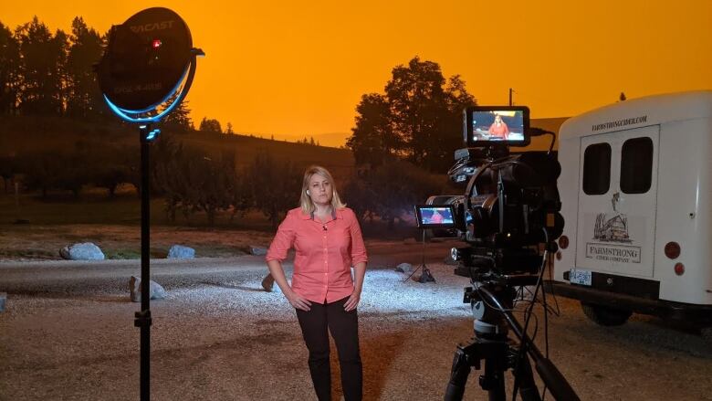 A woman stands in front of a bright orange sky, facing a television camera ready to record