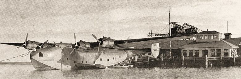 A black and white photo of a sea plane docked next to a wharf with a small building with lots of windows