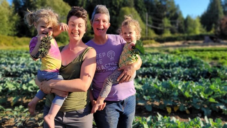 Two women stand smiling in a field of veggies, each holding a toddler. 