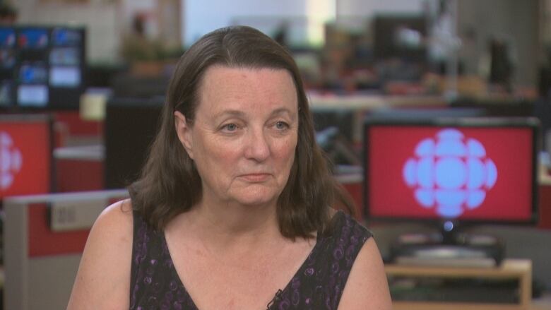 A middle-aged woman with short brown hair sits with the background of the CBC newsroom behind her. 