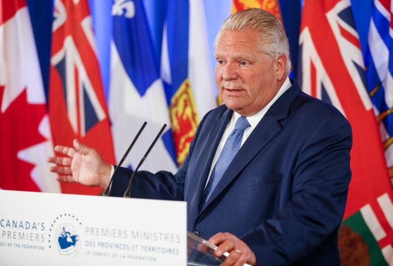 A white man speaks at a podium, with multiple provincial flags behind him.