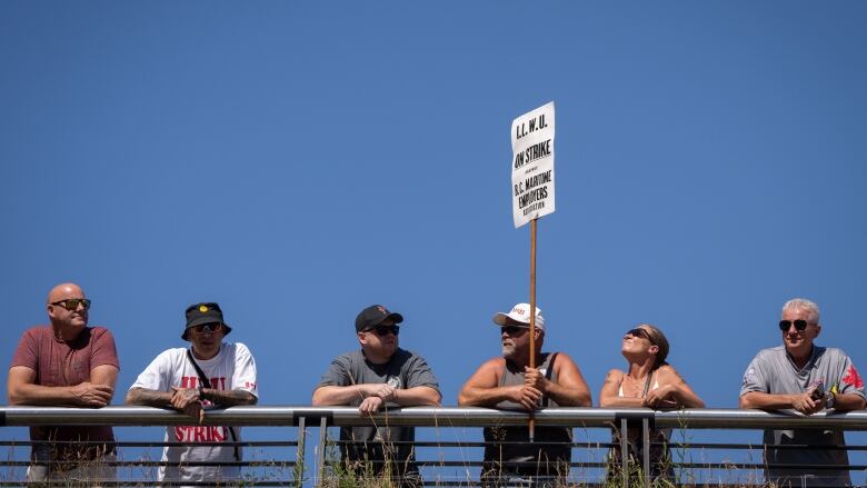 Six people stand shoulder to shoulder at a metal barrier against a blue sky. One man in a grey tank top and white baseball cap holds a white picket sign with the words 