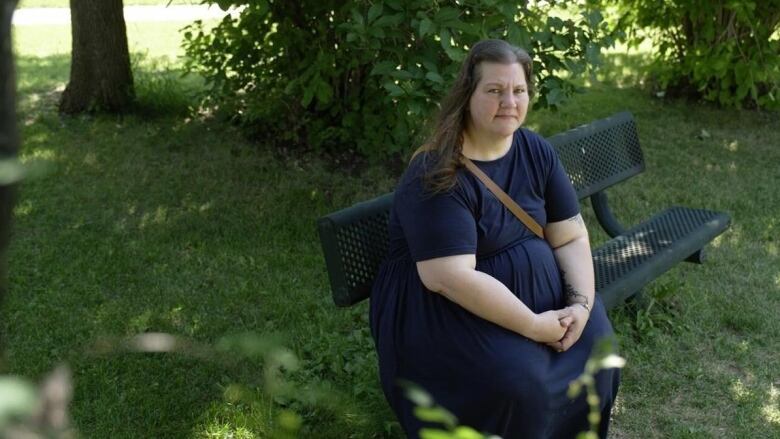 A woman sits on a bench in the shaded area of a park with some trees and a pathway in the background.