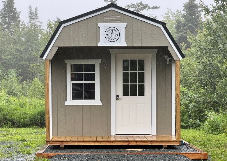 A grey shed with a white door and window in the foreground. Trees and grass in the background.