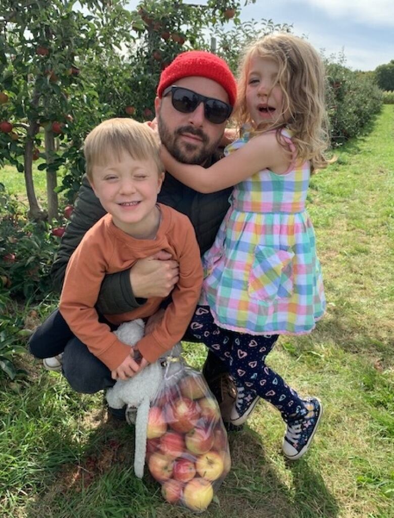 A father and two kids, a young boy and a young girl, in an apple orchard. The son is clutching a bag of apples. 