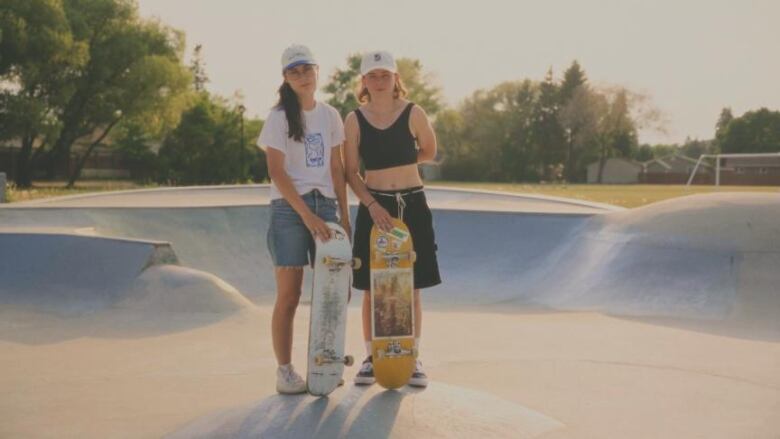 Two skateboarders stand with their boards. One has long brown hair, the other one has shorter hair and is wearing a baseball cap. 