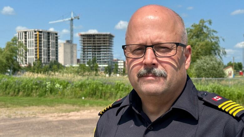 A bald man in a black shirt with his name and a Canada flag in a dirt parking lot with greenery behind him as well as two 15 storey towers under construction. 