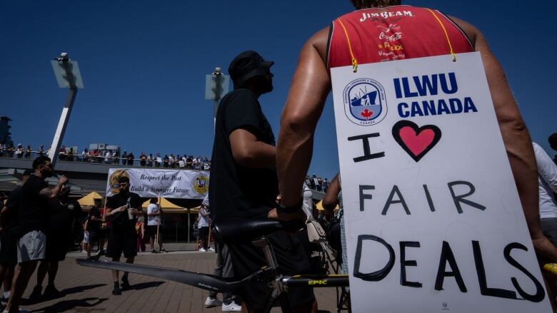 A dock worker is pictured from behind wearing a sign down their back made of cardboard that is tied by a rope to their neck. The sign says I heart fair deals.