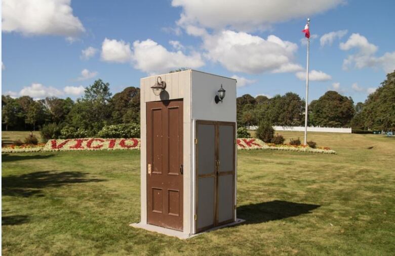 A rectangular kiosk sits on the grass of a park, with each side only slightly bigger than the door it contains. All four sides have doors.